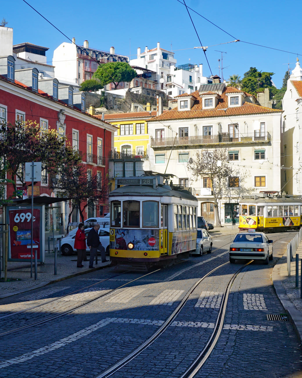 Rua de Lisboa com bondes amarelos, casas coloridas e o bairro de Alfama no alto